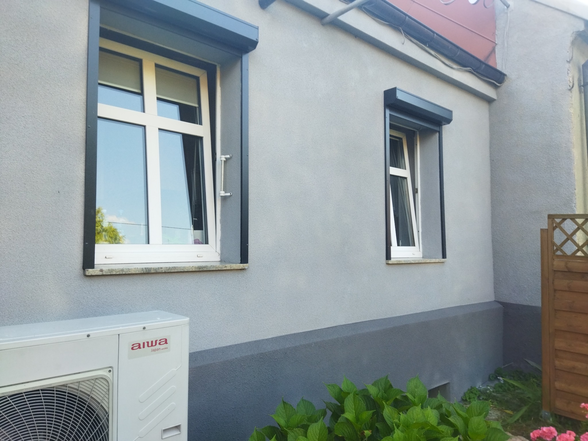 Szara elewacja domu z dwoma oknami z roletami zewnętrznymi, widoczny klimatyzator i fragment drewnianego płotu.