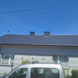 Dom jednorodzinny z ciemnoszarym dachem z dachówki, widoczny zza srebrnego samochodu. Na dachu dwa kominy i antena. Jasne, błękitne niebo.