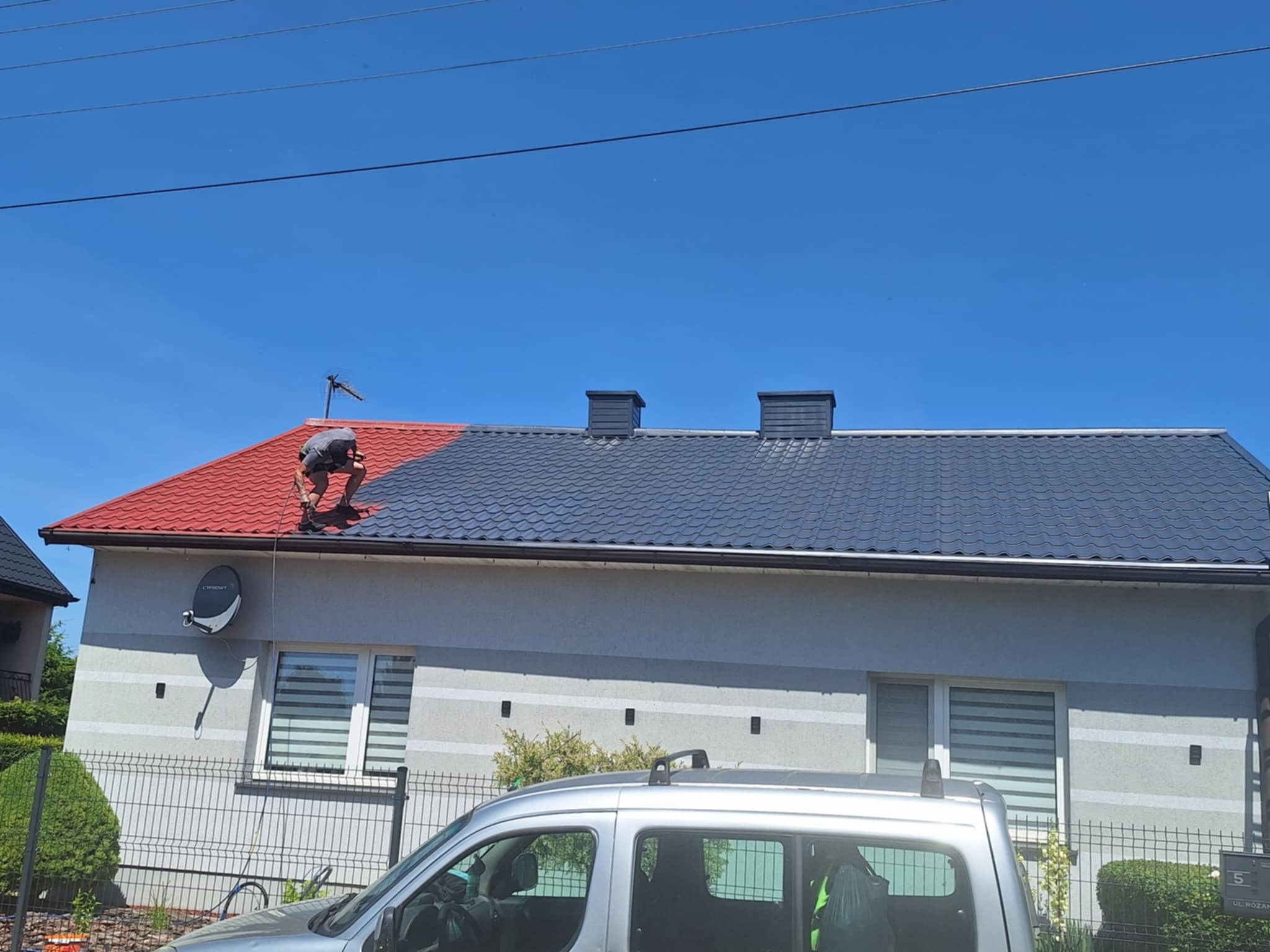 Malowanie dachu domu: część czerwona, część grafitowa. Mężczyzna na dachu w trakcie pracy. Widoczny fragment samochodu i elewacja budynku.