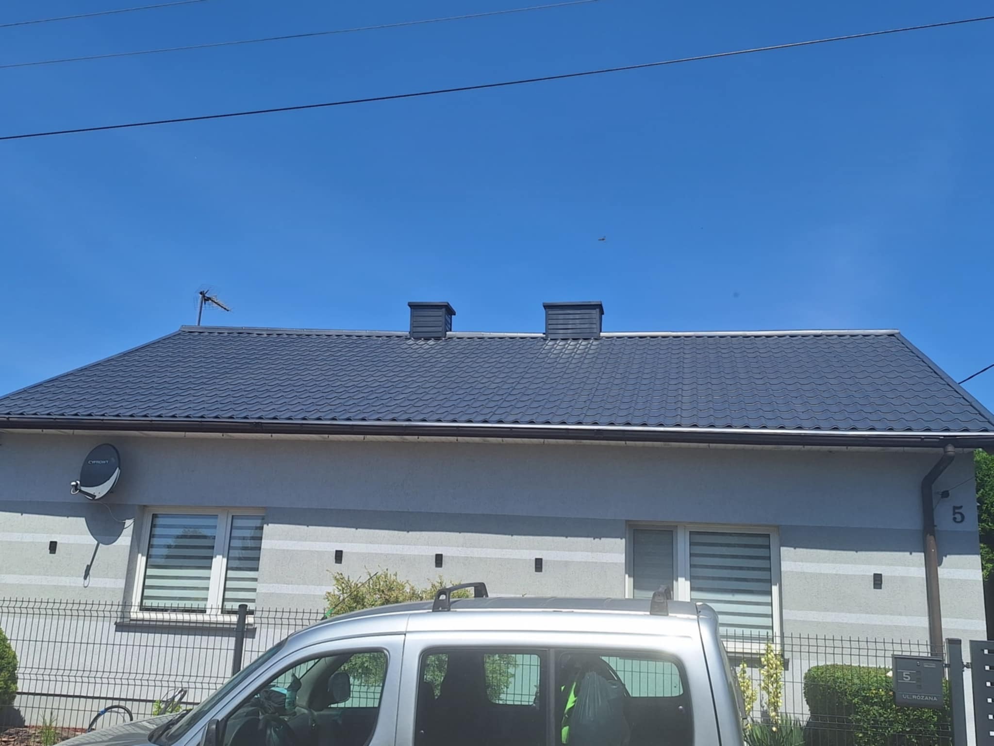 Dom jednorodzinny z ciemnoszarym dachem z dachówki, widoczny zza srebrnego samochodu. Na dachu dwa kominy i antena. Jasne, błękitne niebo.
