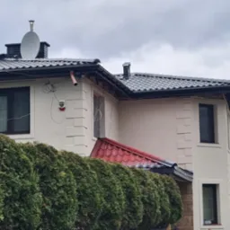 Dwupiętrowy dom z ciemnym dachem, rynnami i satelitą, częściowo zasłonięty żywopłotem, widok na elewację z dekoracyjnymi elementami i wystającymi rurami.