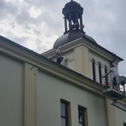Arkadiusz Waśkiewicz - Pracownicy na podnośniku koszowym czyszczą dach i rynny wysokiego budynku z wieżą zwieńczoną krzyżem, widoczne okna z ciemnymi szybami.