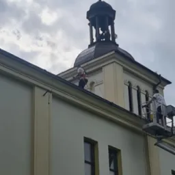 Pracownicy na podnośniku koszowym czyszczą dach i rynny wysokiego budynku z wieżą zwieńczoną krzyżem, widoczne okna z ciemnymi szybami.