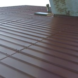 Arkadiusz Waśkiewicz - Ciemnobrązowy dach z blachy trapezowej, widoczny komin z zaciekami i fragment okna dachowego, w tle drzewa i budynek.