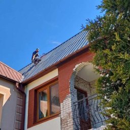 Arkadiusz Waśkiewicz - Pracownik w kasku zabezpieczającym na dachu dwupiętrowego domu z ceglaną elewacją, montujący blachę trapezową na tle błękitnego nieba.