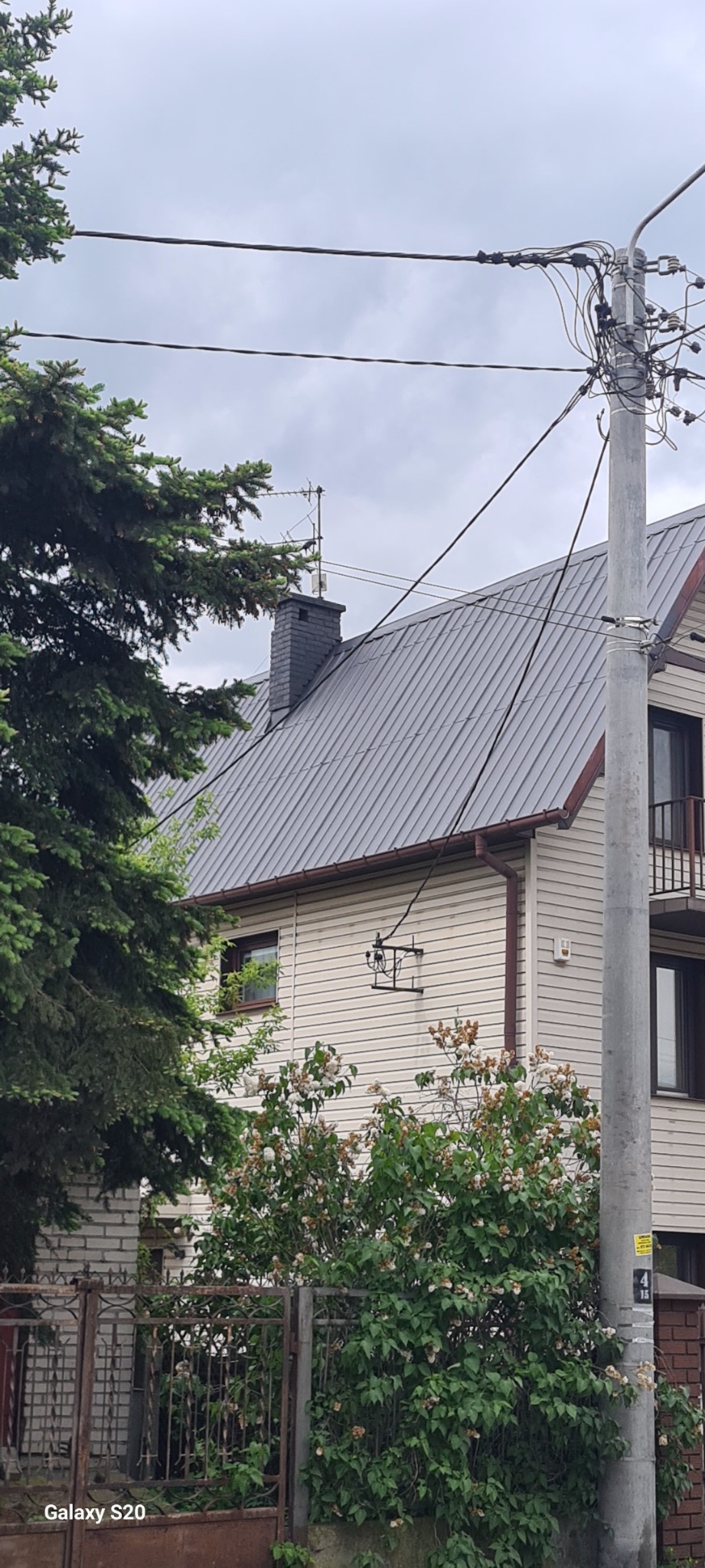 Dwupiętrowy dom z szarą blachą na dachu, widoczny komin, okno, rynna, fragment elewacji z drewnianą okładziną oraz krzewy przed ogrodzeniem.