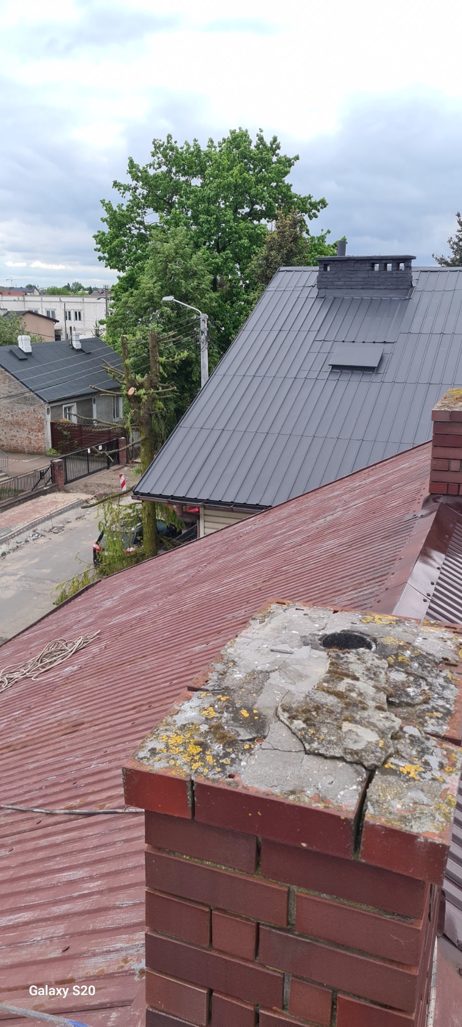 Widok z góry na czerwony, skorodowany dach z ceglanym kominem pokrytym mchem i pęknięciami, w tle szary dach sąsiedniego budynku i zieleń drzew.