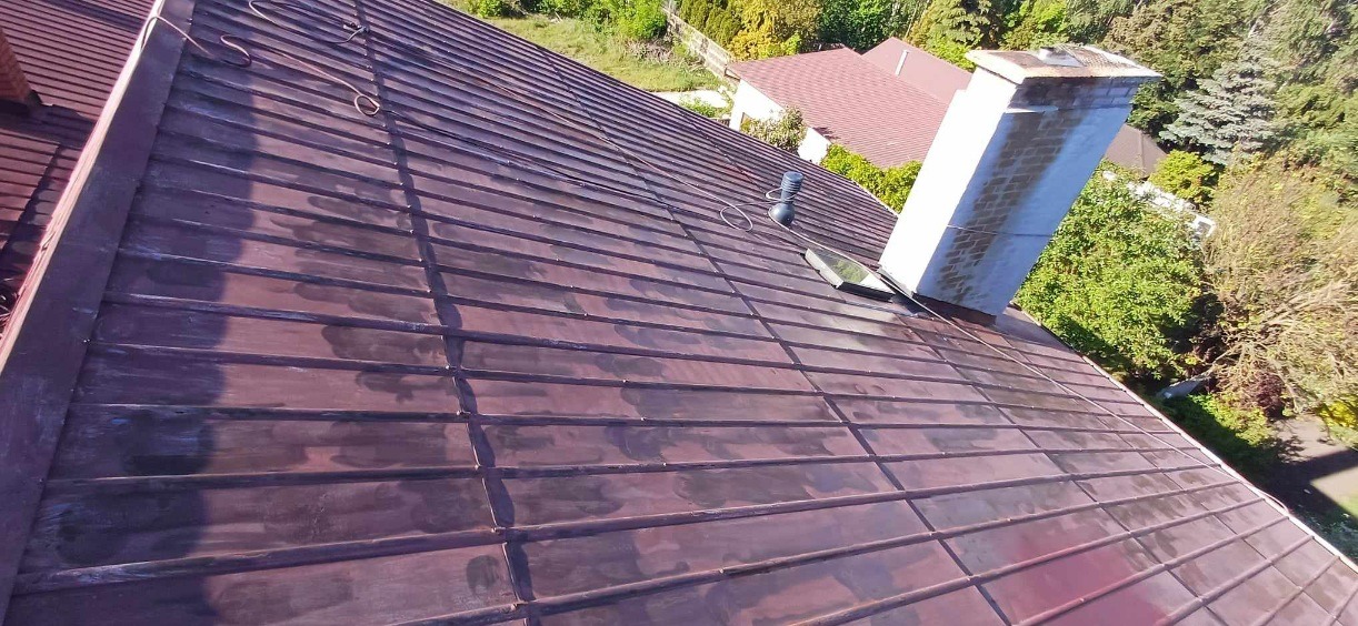 Metalowy dach z kominem i oknem dachowym, widoczne ślady po myciu, na tle zieleni.