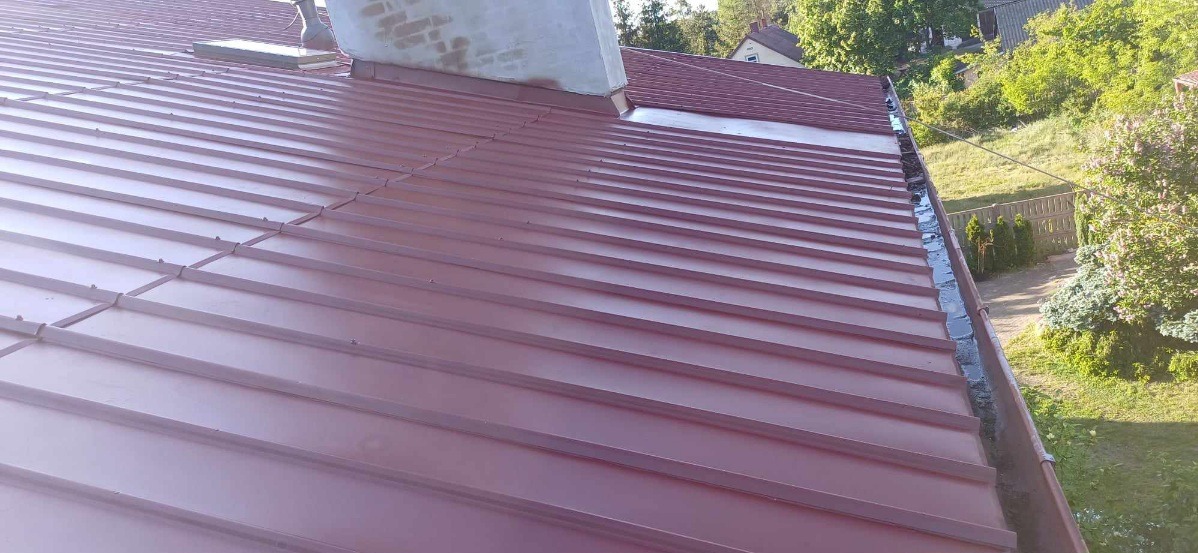 Czerwony dach z blachy trapezowej, widoczny komin i rynna, w tle zieleń drzew i krzewów.