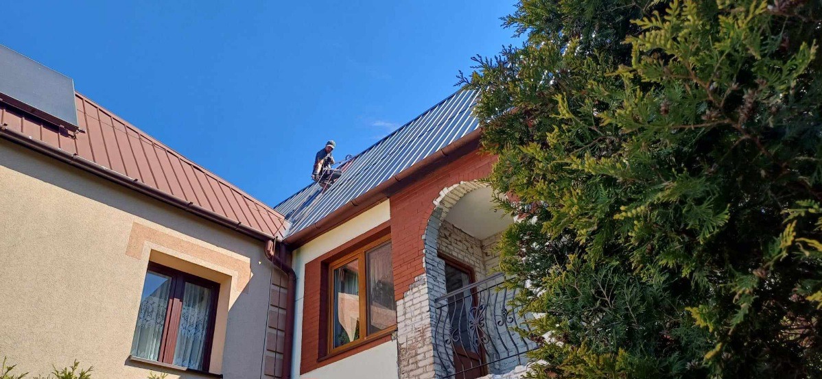 Pracownik w kasku zabezpieczającym na dachu dwupiętrowego domu z ceglaną elewacją, montujący blachę trapezową na tle błękitnego nieba.