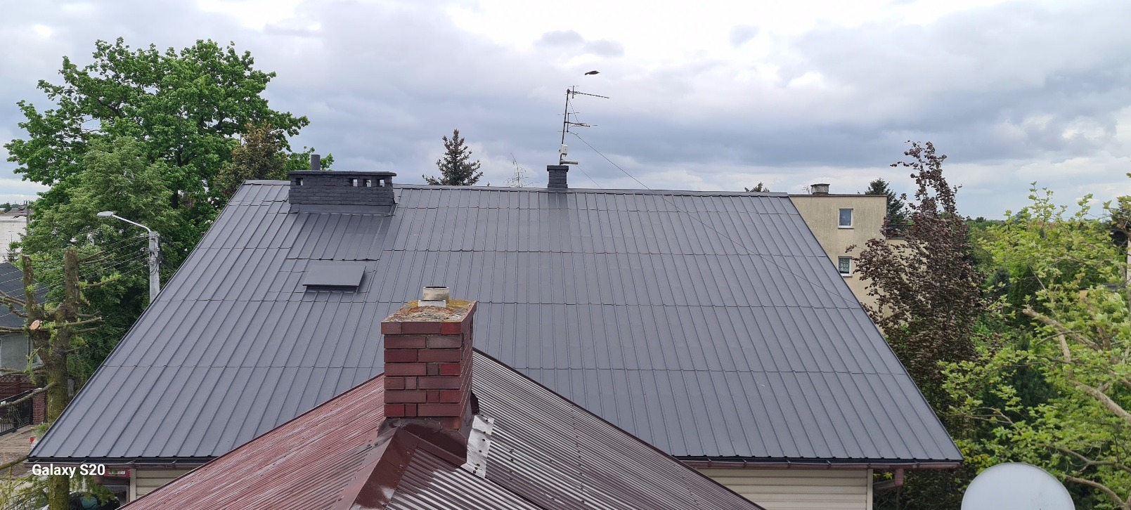 Widok z góry na szary dach z blachy trapezowej z dwoma kominami, anteną i fragmentem dachu pokrytego czerwoną blachą, otoczony zielenią drzew i budynkami mieszkalnymi w pochmurny dzień.