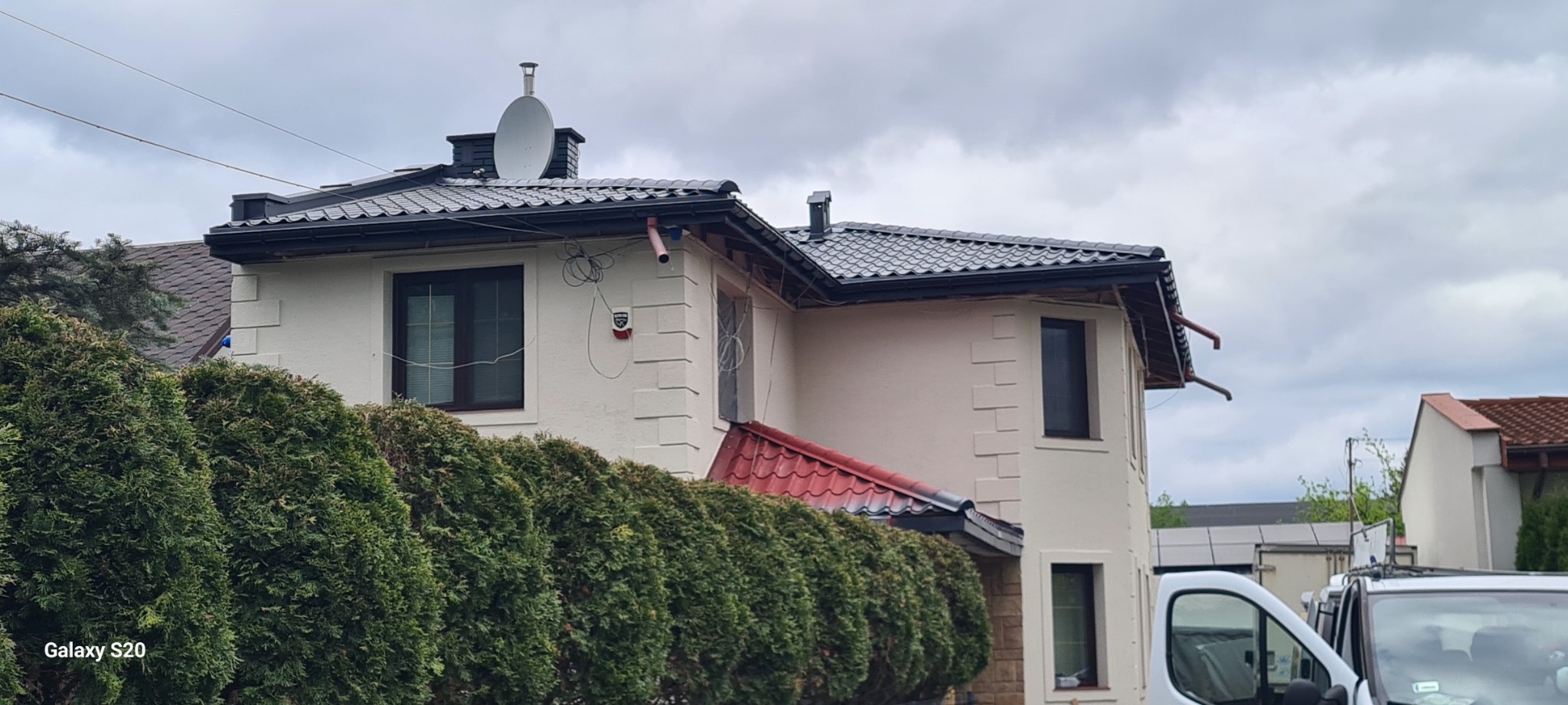 Dom z nowym, ciemnym dachem i rynnami, widoczne także elementy elewacji i antena satelitarna, na pierwszym planie rząd tuj.