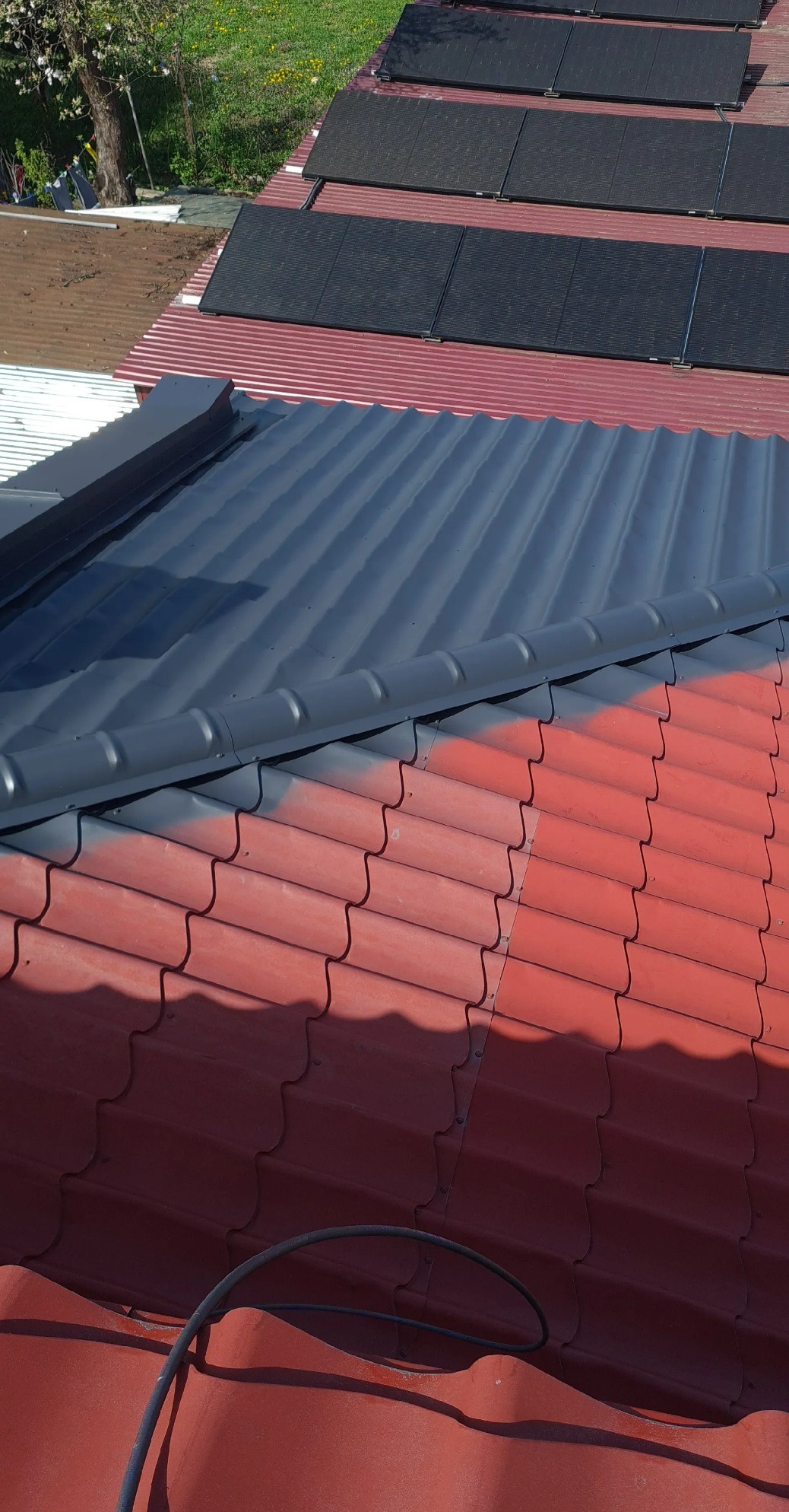 Częściowo pomalowany dach z czerwonej dachówki na szaro, widoczne panele słoneczne w tle i fragment węża na dachu.