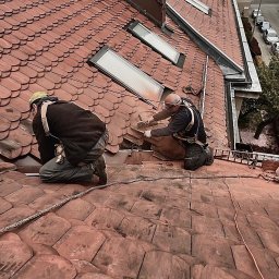 Dach-Art - Dwóch dekarzy zabezpieczonych linami pracuje na dachu krytym czerwoną dachówką, jeden z nich przycina dachówkę szlifierką kątową w pobliżu okien dachowych.