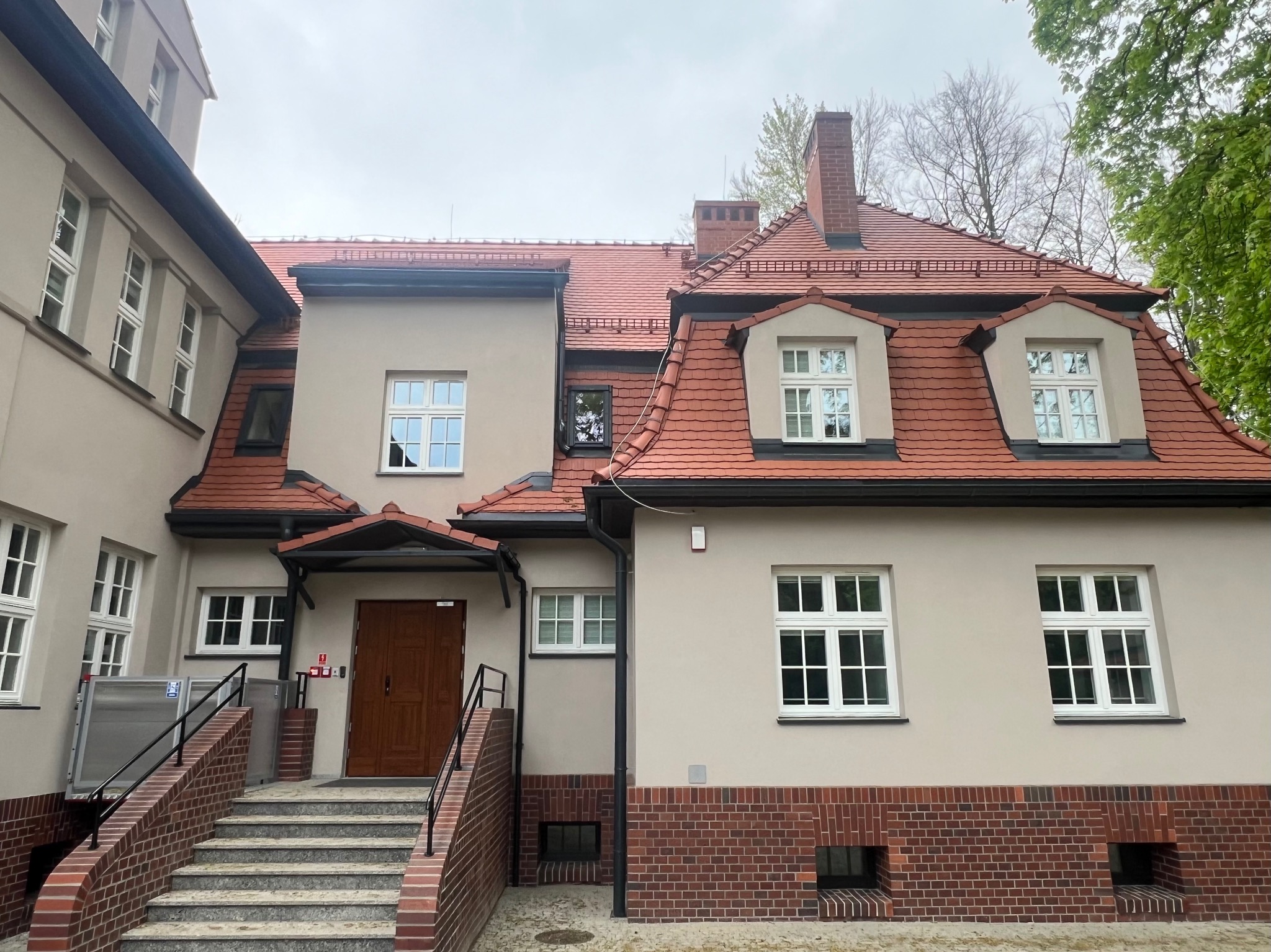 Odnowiony budynek z czerwoną dachówką, widoczne okna dachowe, kominy i schody wejściowe z podjazdem dla niepełnosprawnych.