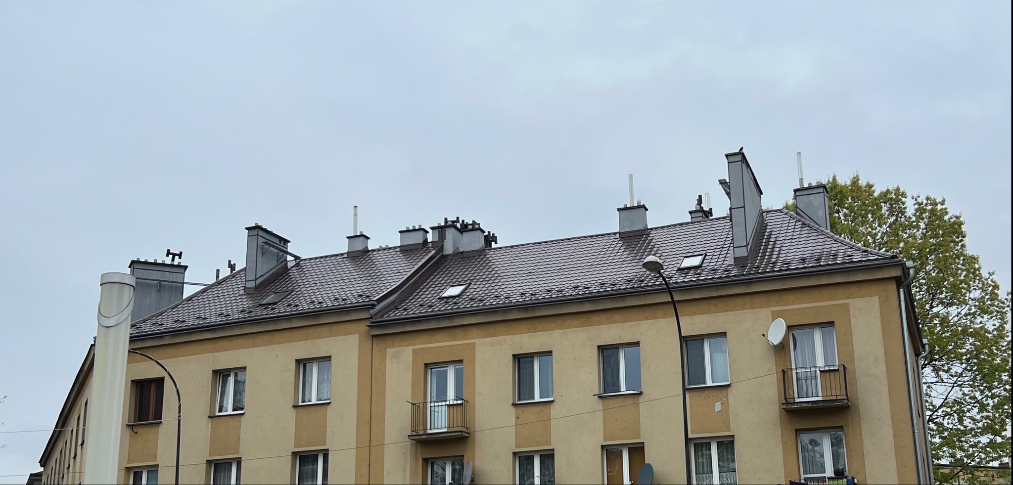 Szary dach z dachówki karpiówki na dwupiętrowym budynku z widocznymi kominami, lukarnami i antenami satelitarnymi.