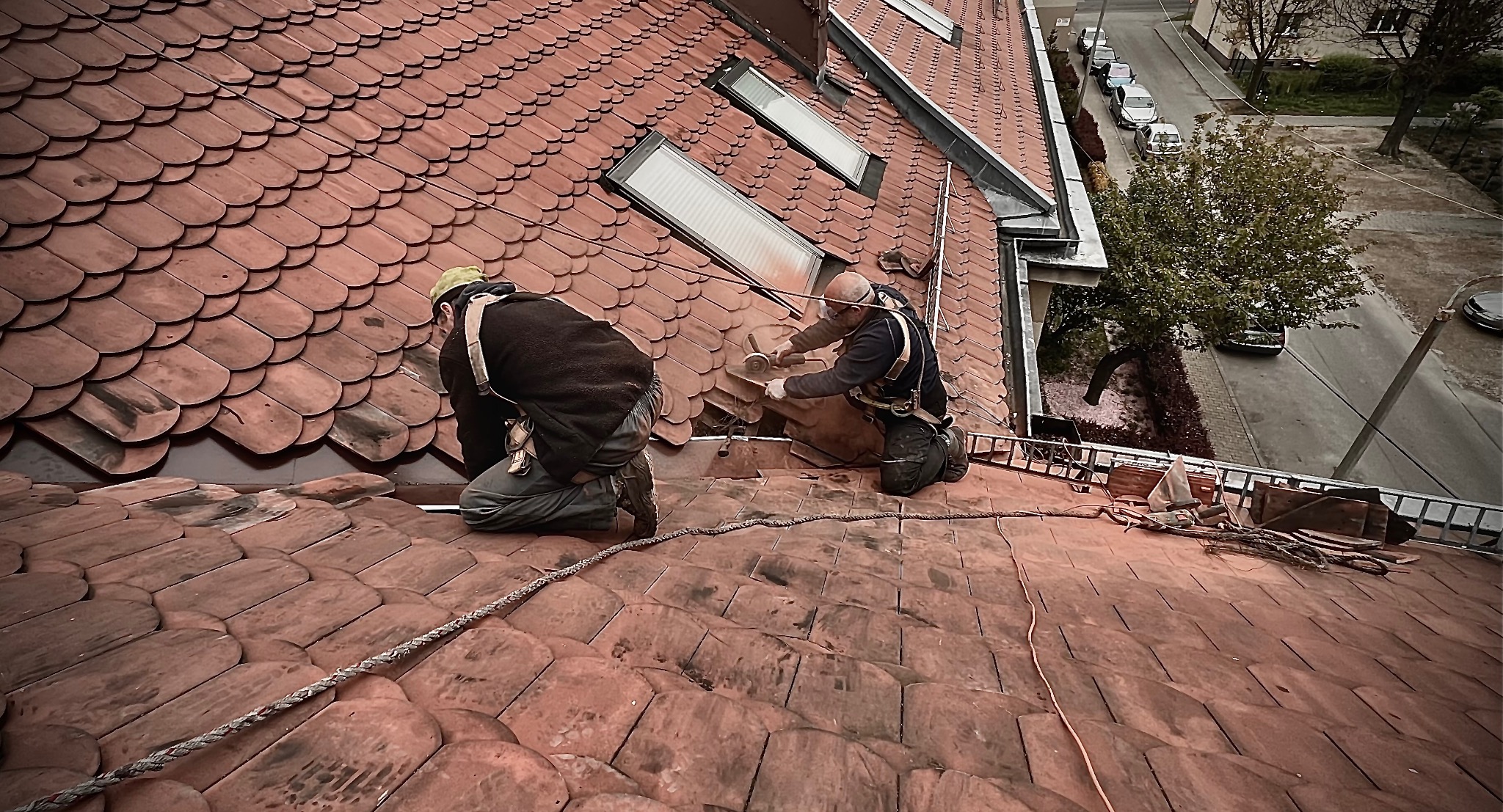 Dwóch dekarzy zabezpieczonych linami pracuje na dachu krytym czerwoną dachówką, jeden z nich przycina dachówkę szlifierką kątową w pobliżu okien dachowych.