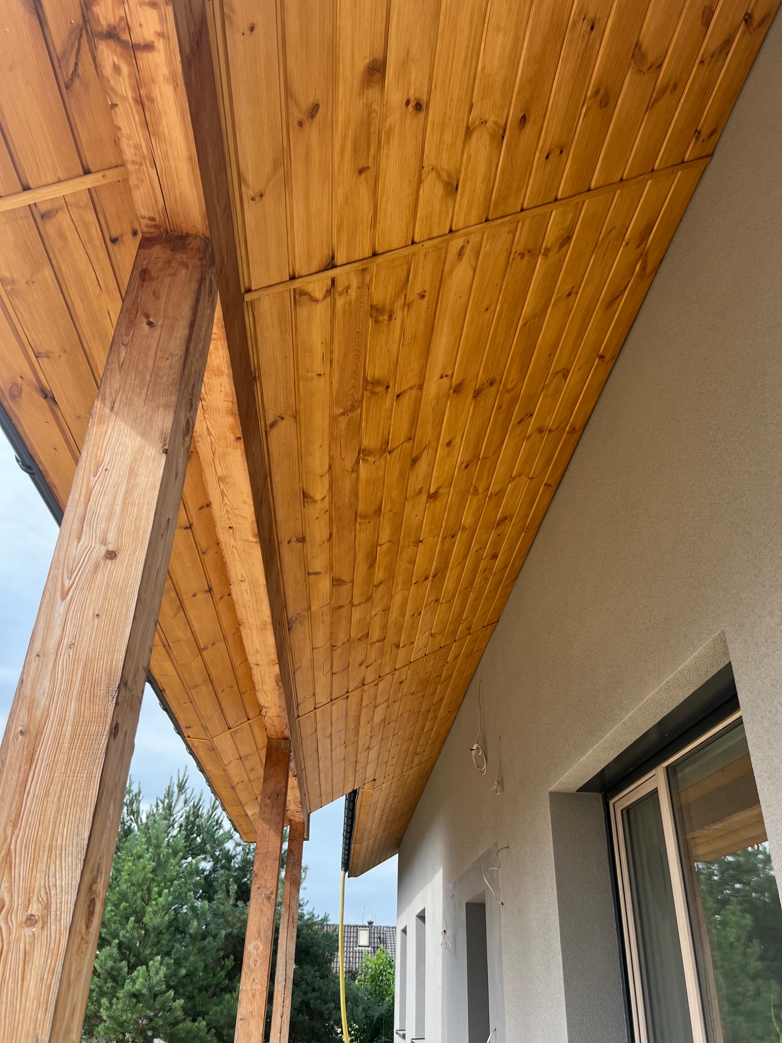 Drewniana podbitka dachowa z widocznymi belkami nośnymi, świeżo zamontowana przy elewacji budynku w trakcie wykańczania.