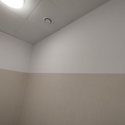 GROTBUD - Pomieszczenie z dwukolorową ścianą: górna część biała, dolna beżowa, oraz biały sufit z okrągłą kratką wentylacyjną i lampą.