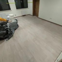 Pokój po ułożeniu jasnych paneli podłogowych; widoczne okna, grzejnik i drzwi; na podłodze leżą pędzle, wiaderka z farbą i folia ochronna.