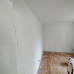 GROTBUD - Pokój po odświeżeniu ścian na biało, widoczne ślady po taśmie malarskiej na styku ściany i sufitu, oraz zabrudzenia na podłodze.