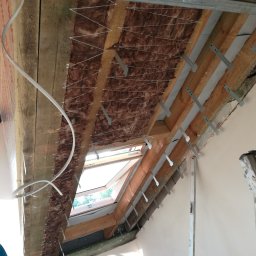 GROTBUD - Montaż izolacji z wełny mineralnej na poddaszu, widoczne drewniane belki, metalowe profile i okno dachowe.