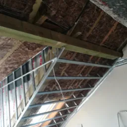 Skośny sufit poddasza w trakcie izolacji wełną mineralną i montażu metalowej konstrukcji pod płyty gipsowo-kartonowe, widoczne okno dachowe i drabina aluminiowa w tle.