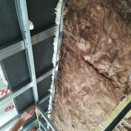 GROTBUD - Montaż sufitu podwieszanego z izolacją z wełny mineralnej, widoczne profile metalowe i ręce mierzące odległość taśmą mierniczą.