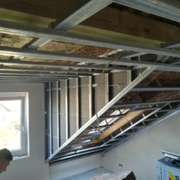 GROTBUD - Montaż metalowego stelaża sufitu podwieszanego z izolacją z wełny mineralnej, widoczny fragment elewacji budynku przez okno i materiały budowlane w rogu pomieszczenia.