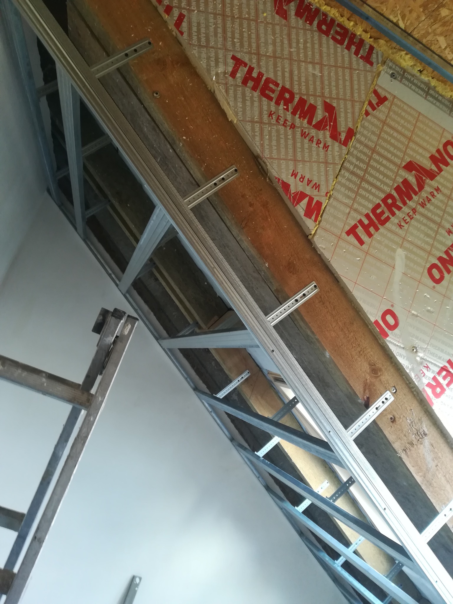 Montaż stelaża z profili stalowych na drewnianej konstrukcji, widoczna izolacja termiczna z nadrukiem 'THERMANO' oraz fragment drabiny malarskiej.
