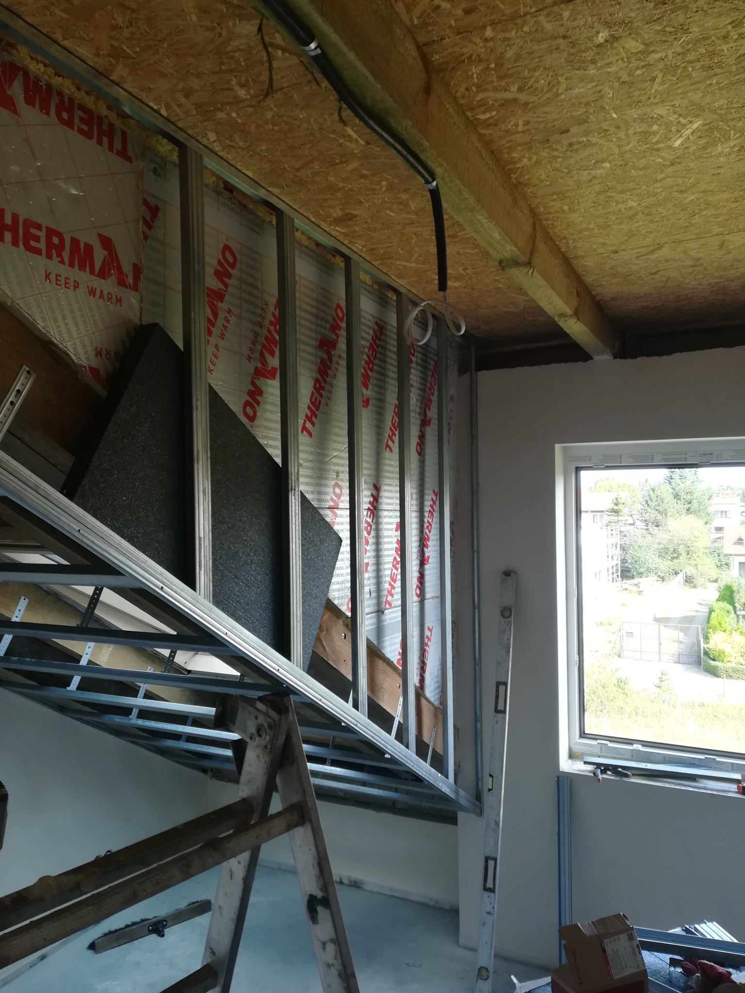 Wnętrze w trakcie wykańczania: widoczne metalowe profile konstrukcyjne, ocieplenie ścian wełną mineralną z folią aluminiową, drewniana podbitka sufitowa, okno z widokiem na zieleń, poziomica oparta...