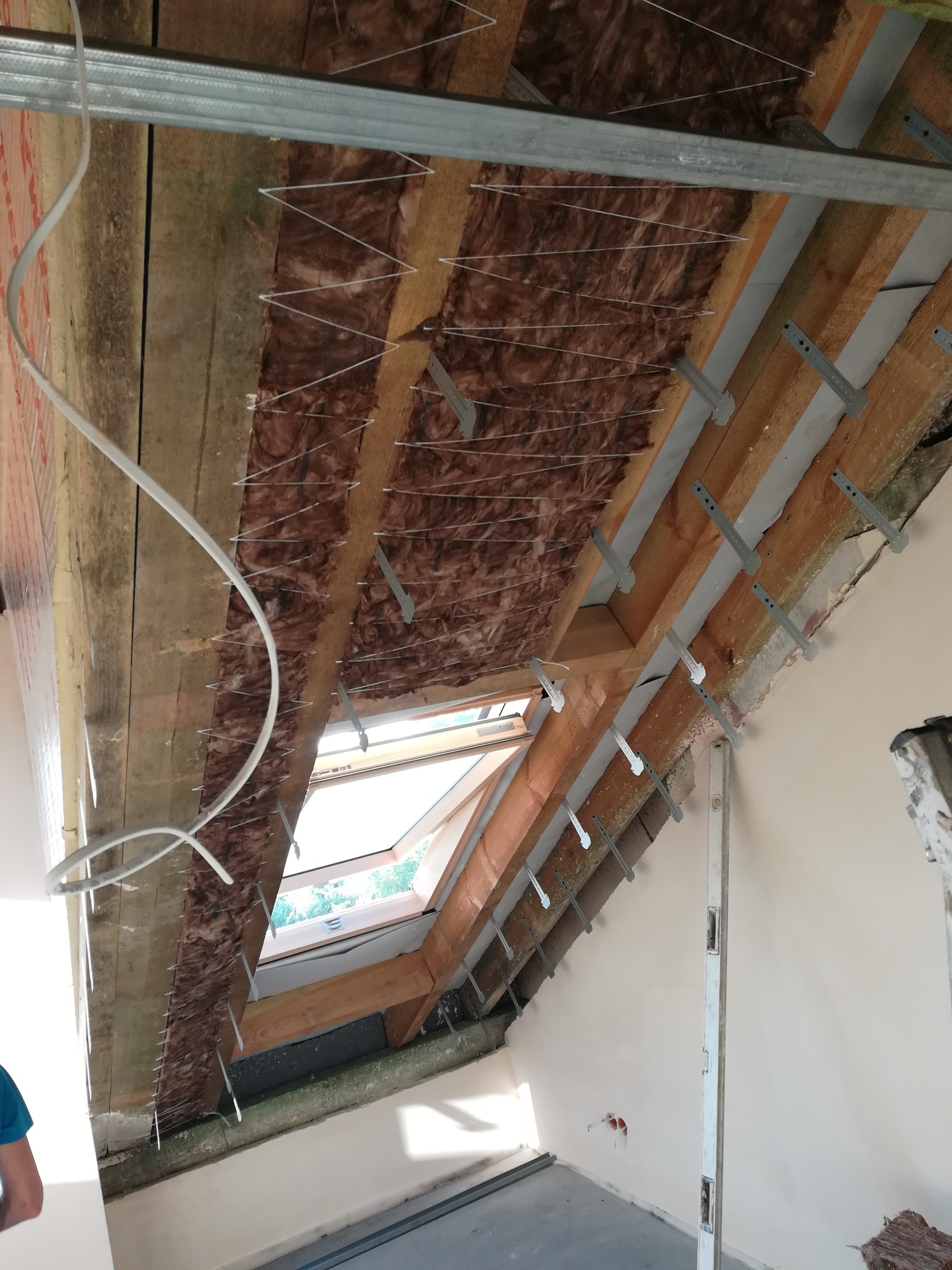 Montaż izolacji z wełny mineralnej na poddaszu, widoczne drewniane belki, metalowe profile i okno dachowe.