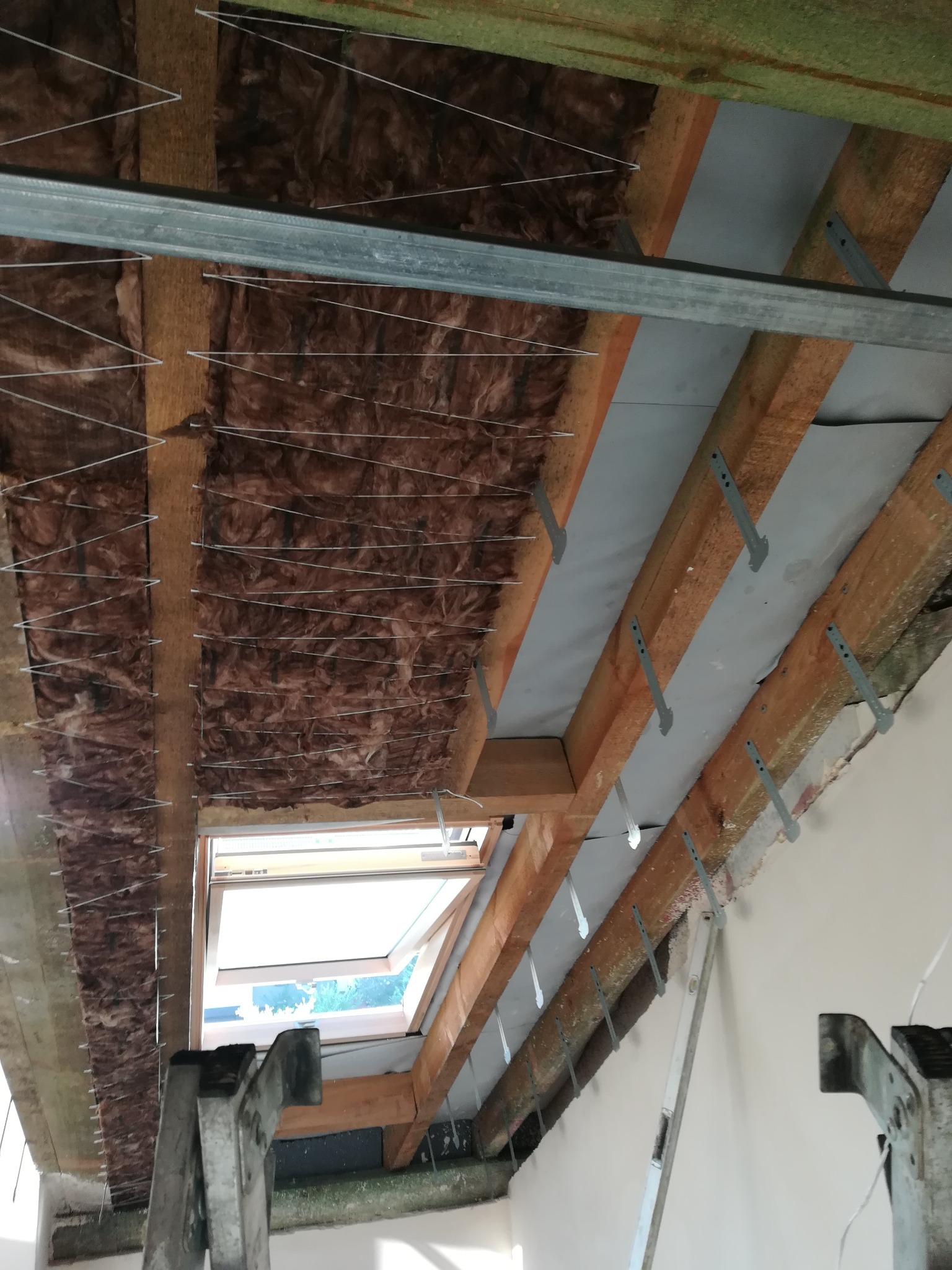 Izolacja poddasza wełną mineralną, widoczne drewniane krokwie, metalowe profile i okno dachowe. Prace w trakcie realizacji.