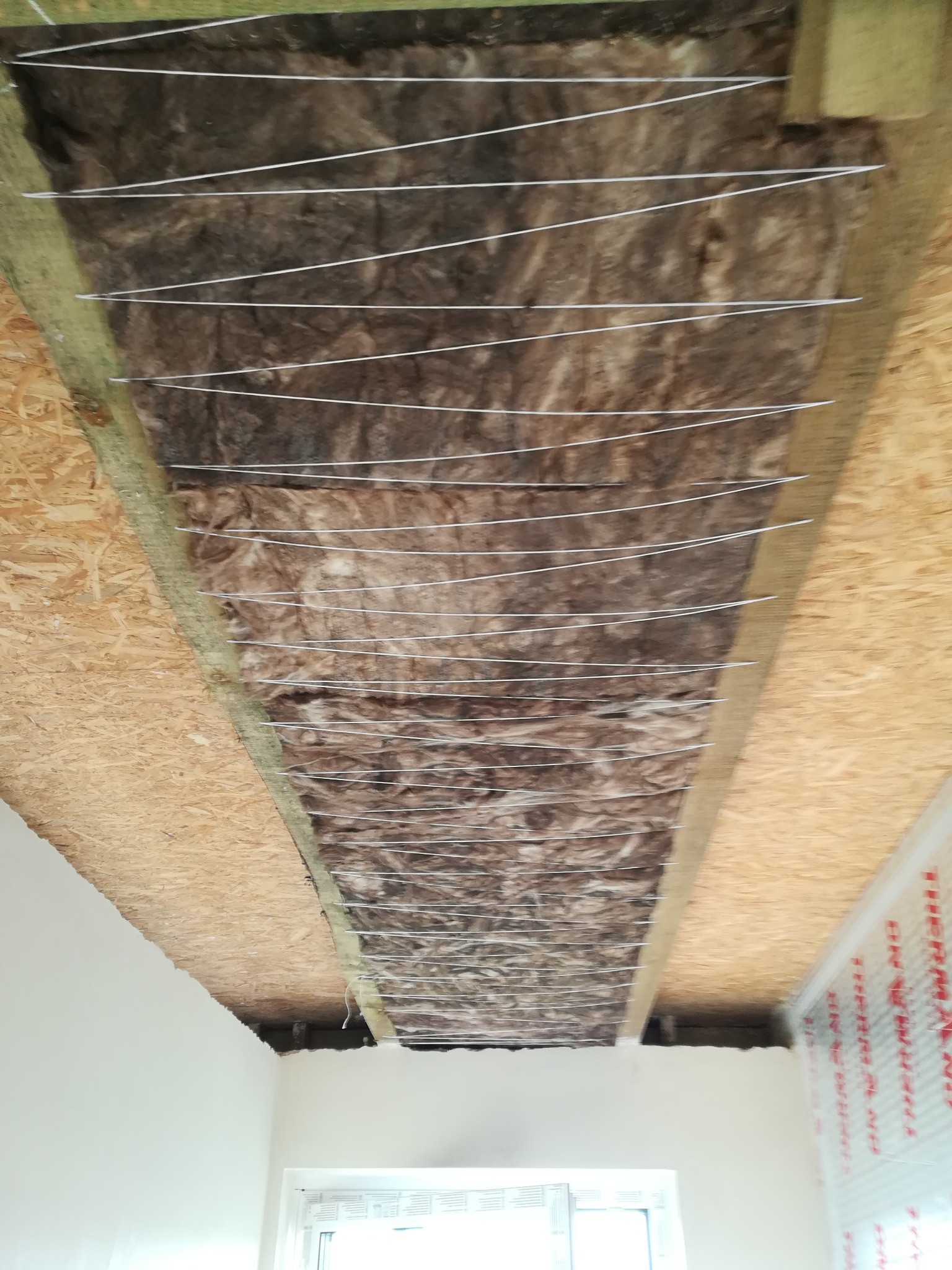 Izolacja termiczna stropu wełną mineralną, zabezpieczona sznurkiem, widoczne elementy drewniane konstrukcji i płyty OSB.