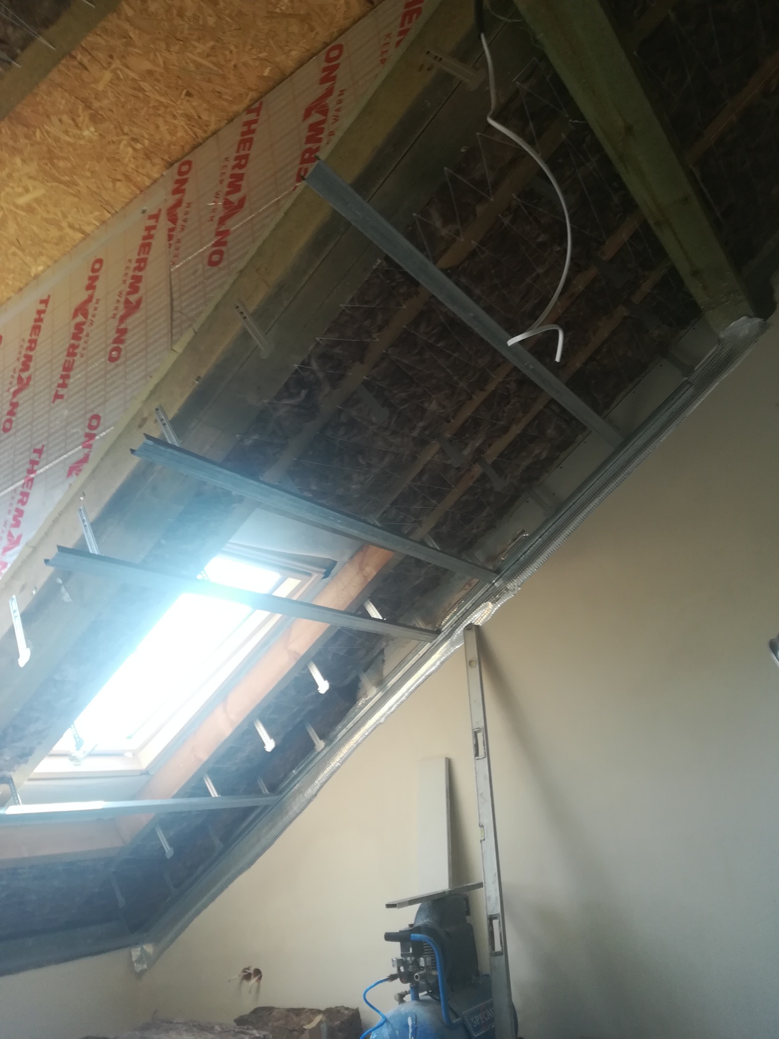 Montaż metalowego stelażu pod płytę gipsowo-kartonową na poddaszu z oknem dachowym, widoczne ocieplenie i fragment kompresora. Etap przygotowawczy do prac wykończeniowych.