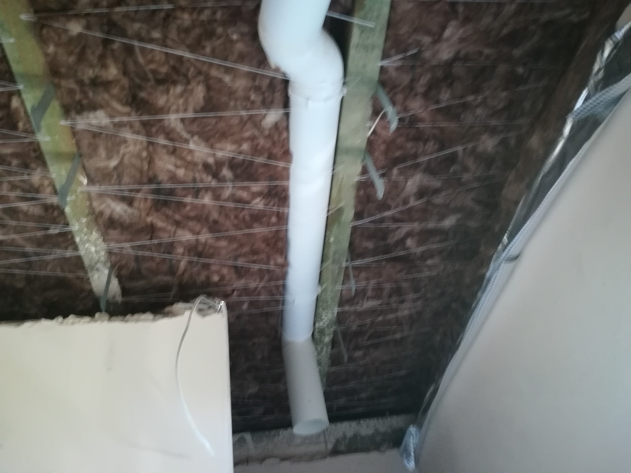 Izolacja poddasza z widoczną rurą wentylacyjną, mocowaną do drewnianej belki za pomocą metalowych uchwytów, oraz fragmentem ściany z płyt gipsowo-kartonowych.