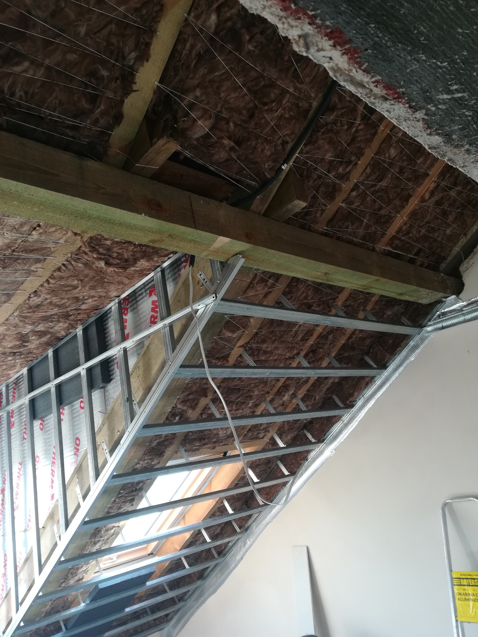 Skośny sufit poddasza w trakcie izolacji wełną mineralną i montażu metalowej konstrukcji pod płyty gipsowo-kartonowe, widoczne okno dachowe i drabina aluminiowa w tle.