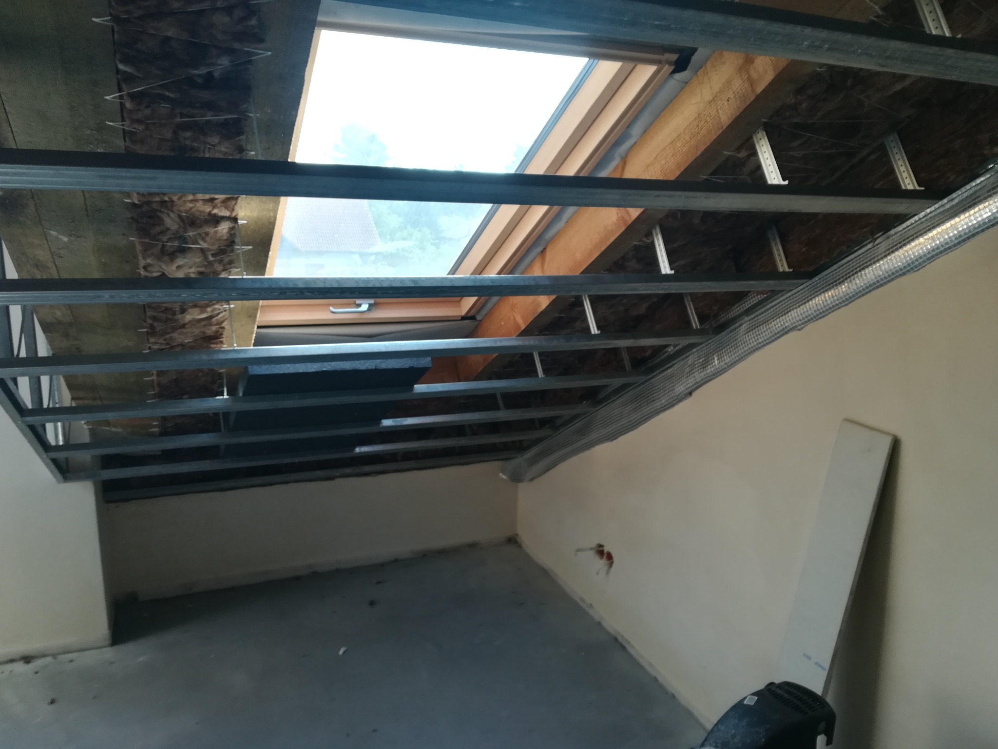 Montaż metalowej konstrukcji sufitu podwieszanego z widocznym oknem dachowym i izolacją, wnętrze w trakcie prac wykończeniowych.