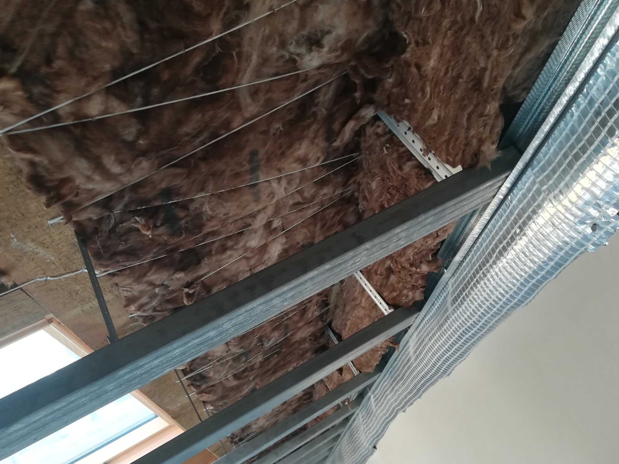 Izolacja termiczna poddasza z widoczną wełną mineralną, metalową konstrukcją nośną i folią paroizolacyjną z siatką wzmacniającą.