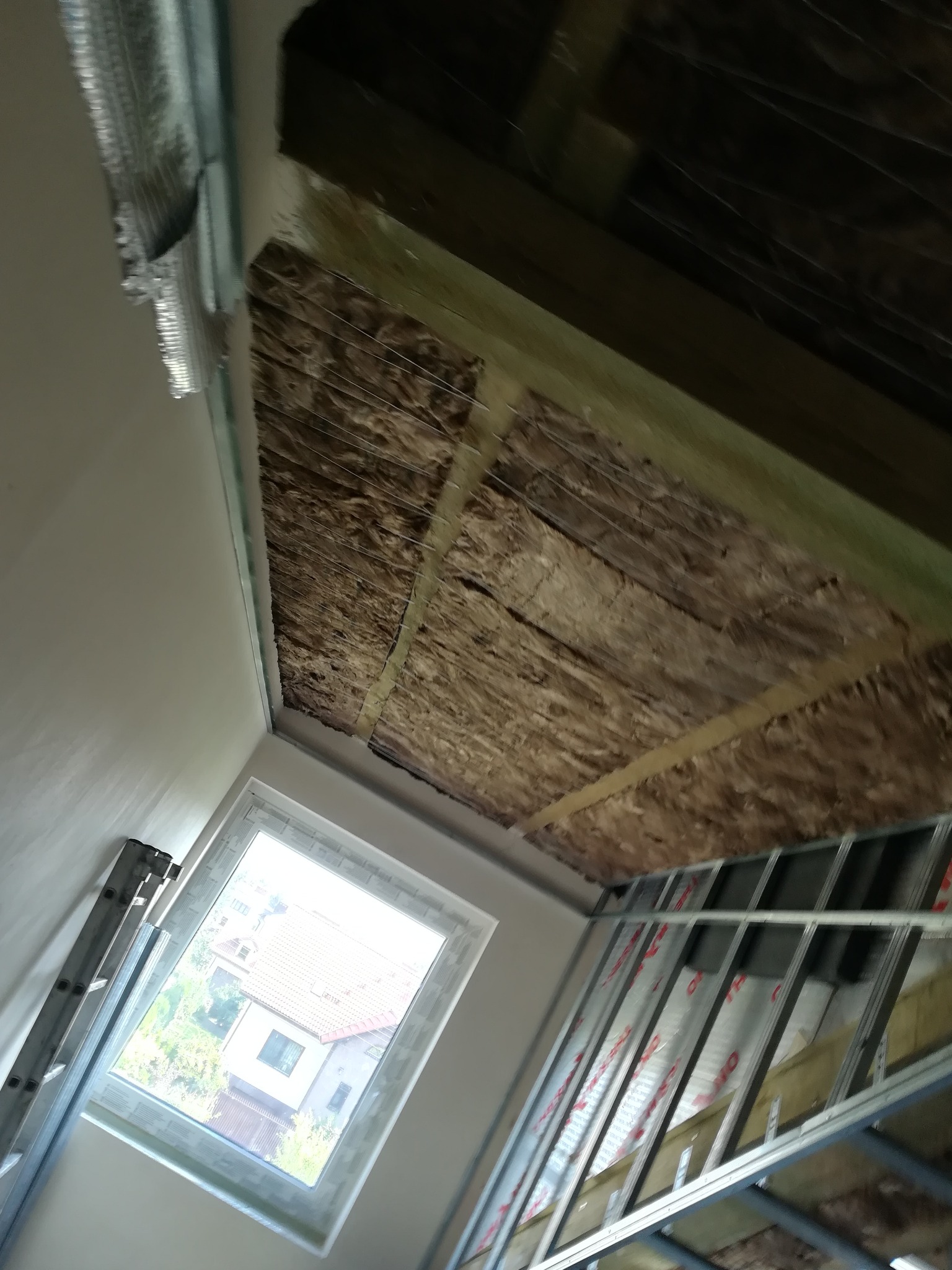 Skośny sufit z widoczną izolacją z wełny mineralnej i metalową konstrukcją pod płytę gipsową, widok na okno dachowe i drabinę opartą o ścianę.
