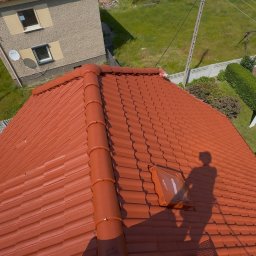 CZYSTAMOC OLEH KUSIE - Czerwony dach z dachówki ceramicznej po renowacji, widoczny cień osoby na dachu i okno dachowe. Widok z góry na dom i trawnik w słoneczny dzień.