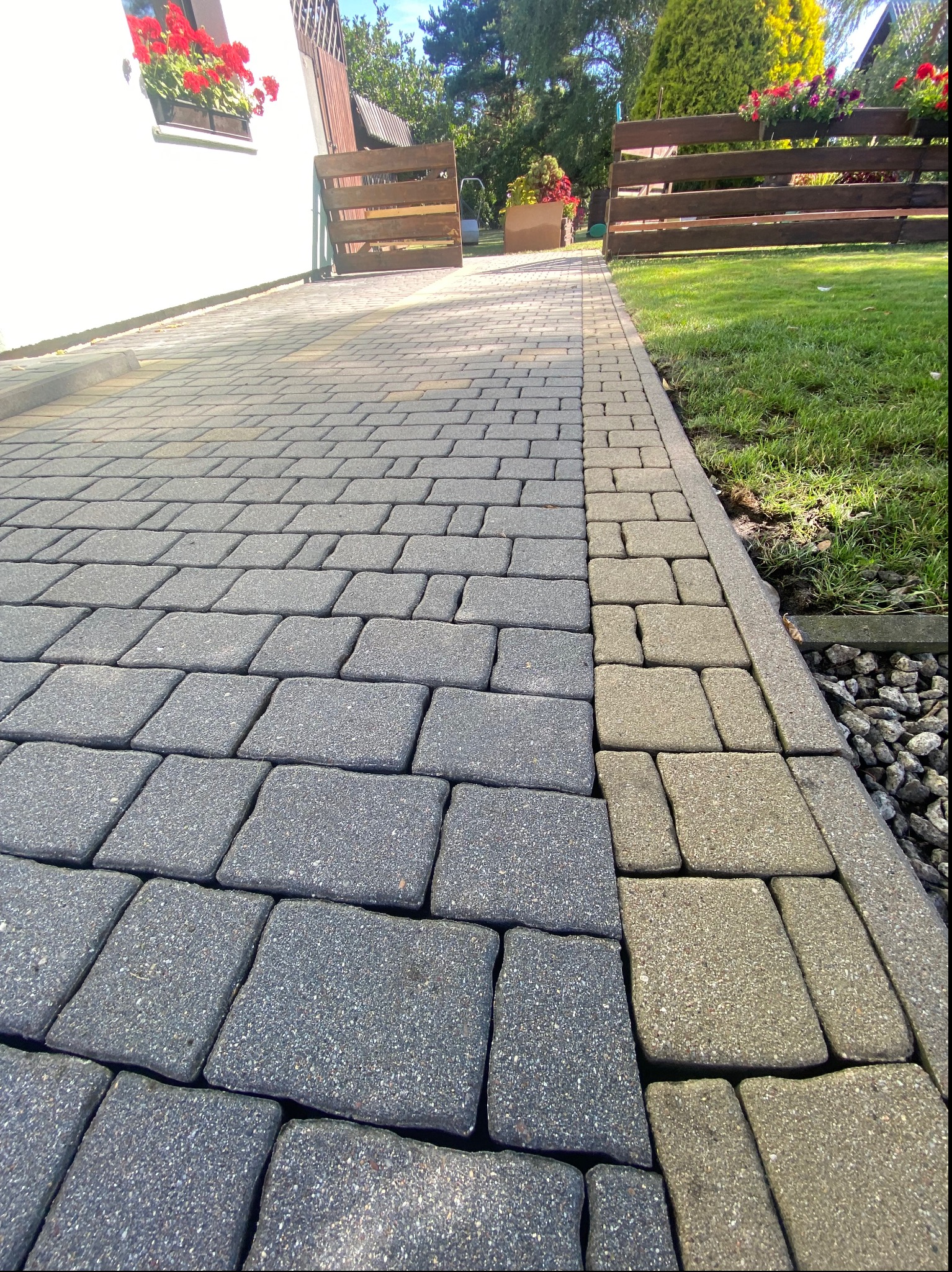 Ułożona szara kostka brukowa na podjeździe z obrzeżem z jaśniejszej kostki, widoczny fragment trawnika i drewniane ogrodzenie z kwiatami.