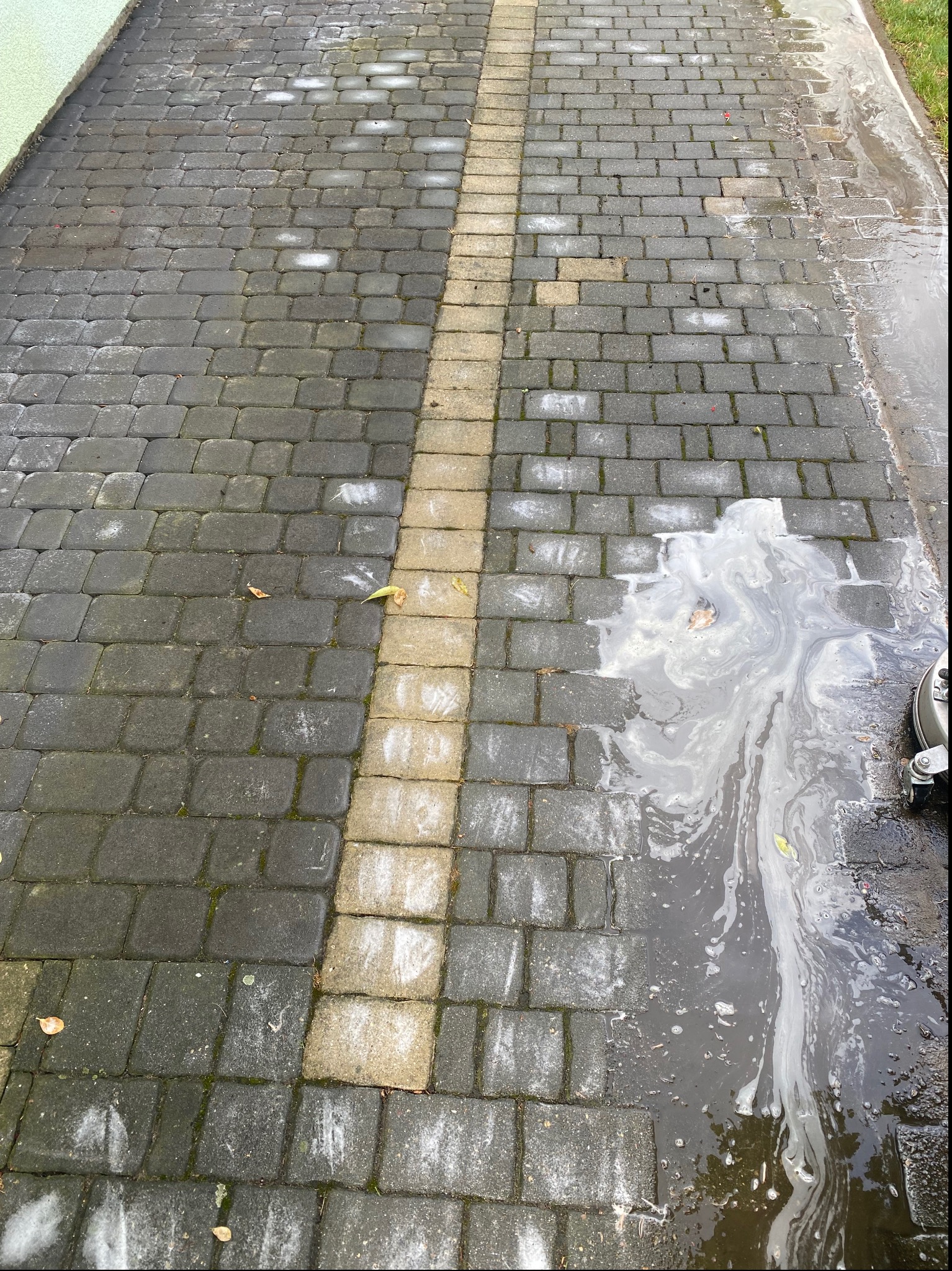 Częściowo umyta kostka brukowa z widocznymi smugami brudnej wody i resztkami liści, z jasnym pasem kostki pośrodku.