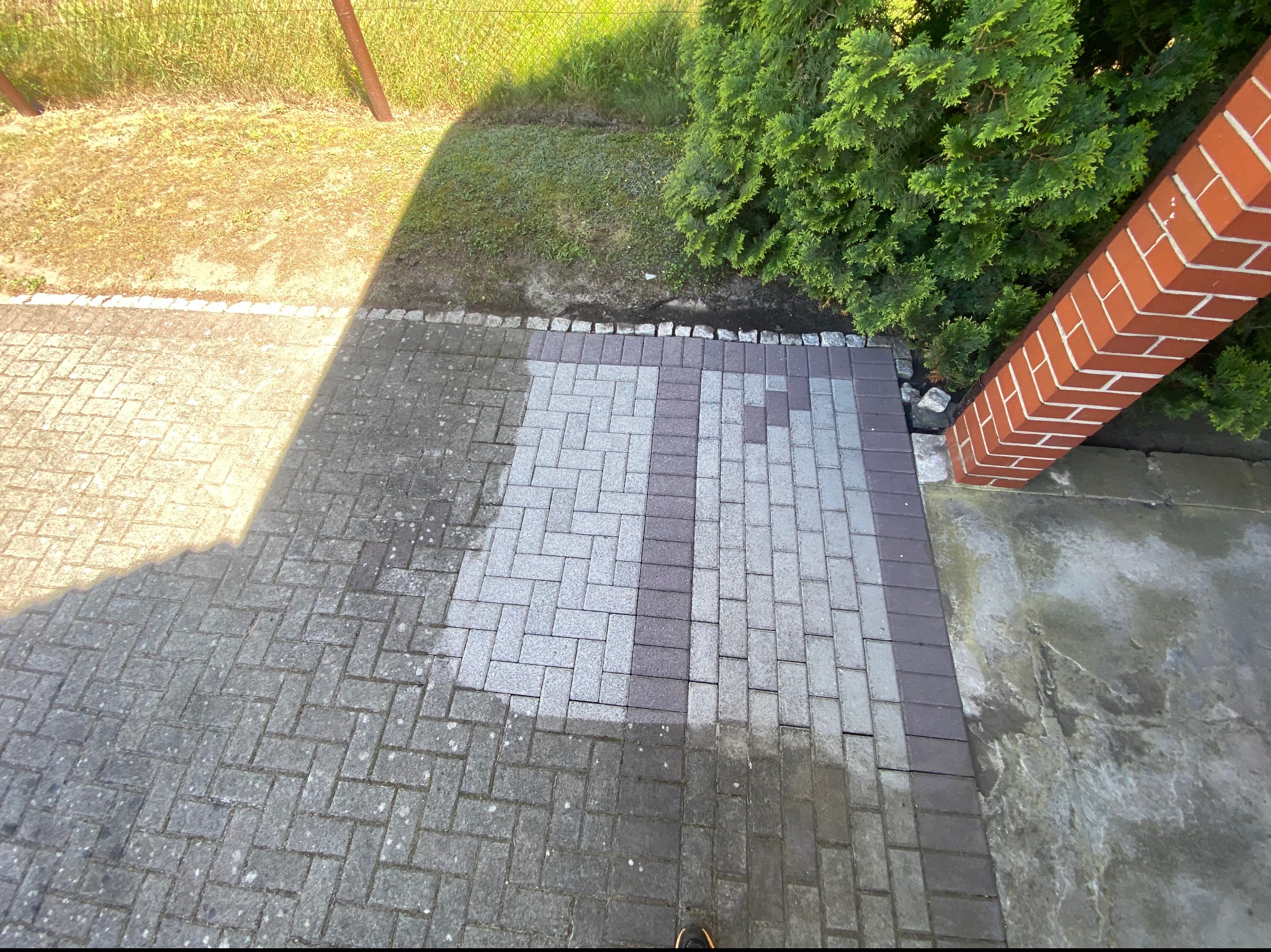 Częściowo wyczyszczona kostka brukowa w odcieniach szarości, z widoczną różnicą w kolorze między czystą a brudną powierzchnią, obok muru z czerwonej cegły i zielonych krzewów.