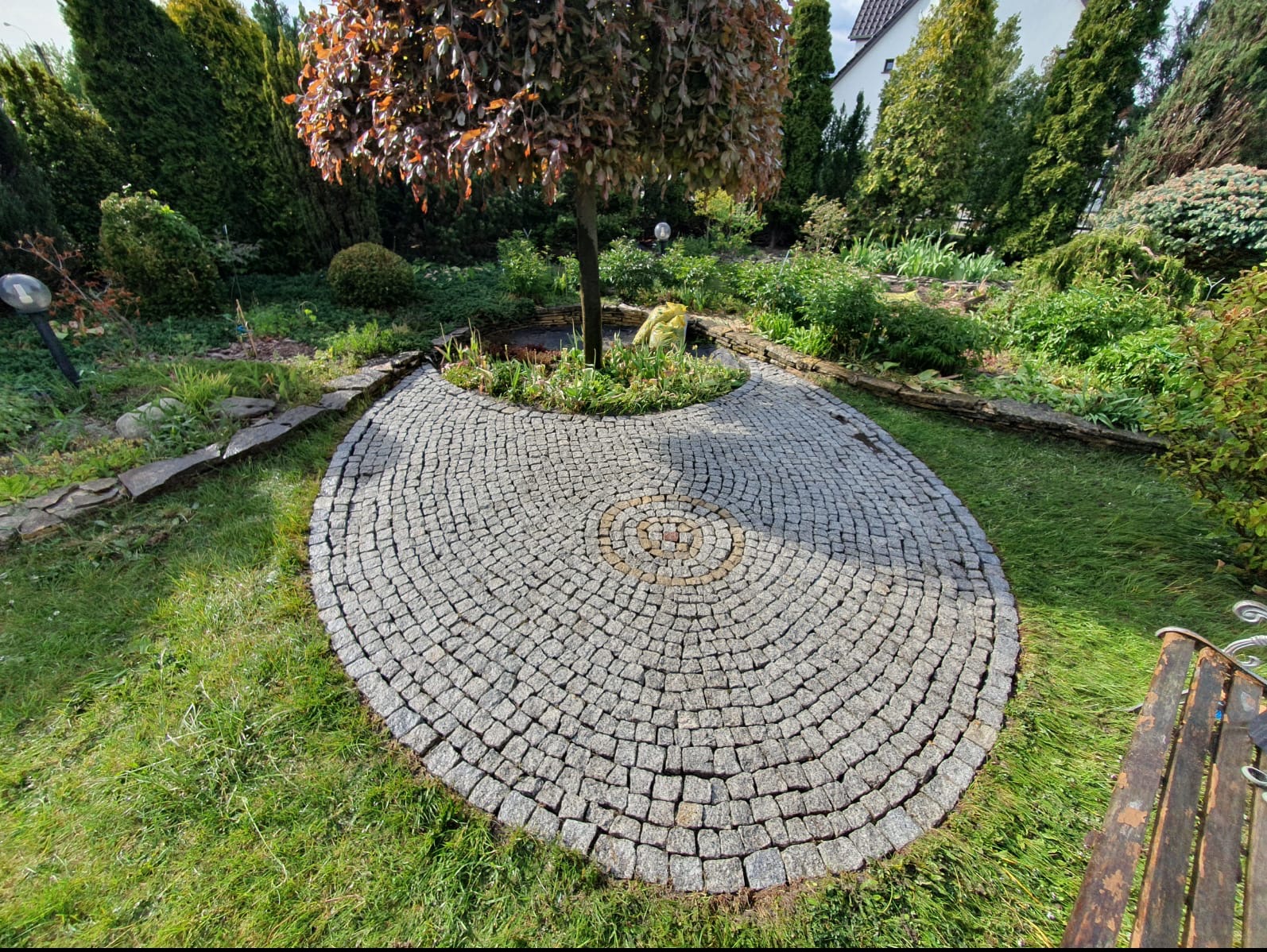 Okrągły plac z kostki brukowej ułożonej w koncentryczne kręgi wokół drzewa w ogrodzie, z centralnym akcentem z jaśniejszych kostek.