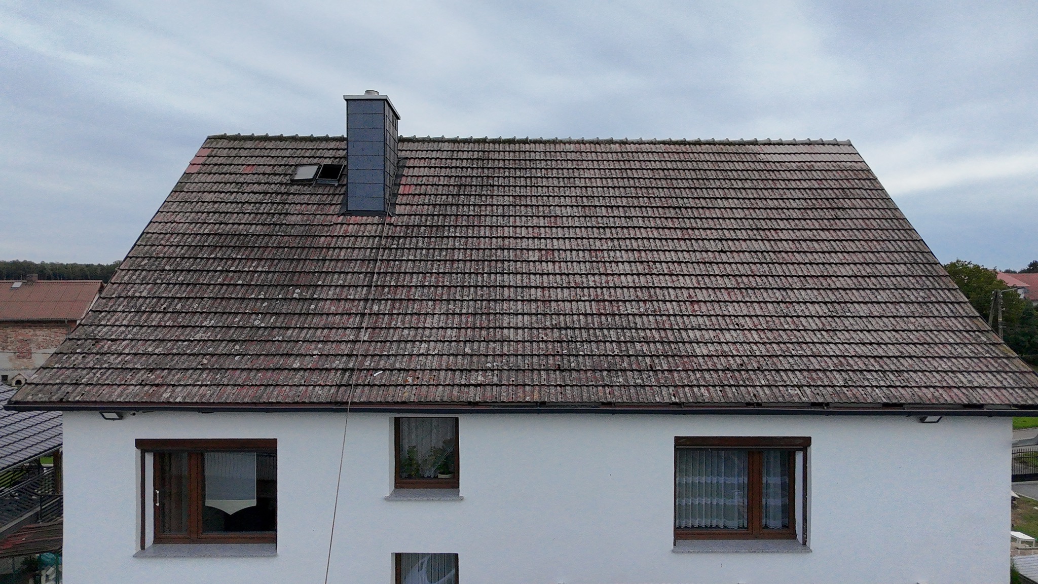 Dom z widocznym, zabrudzonym dachem z dachówki ceramicznej, kominem i oknem dachowym. Biała elewacja budynku z trzema oknami. Pochmurne niebo.