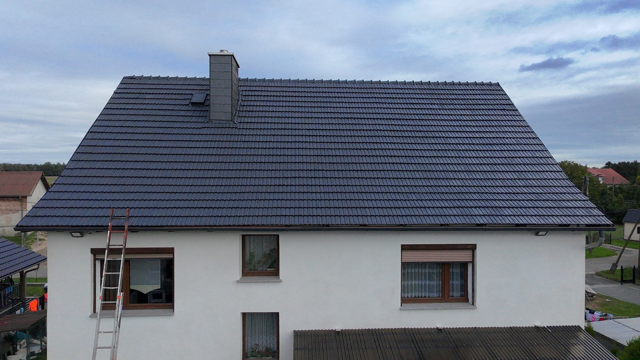 Dom z nowym, ciemnoszarym dachem z dachówki ceramicznej. Metalowa drabina oparta o ścianę. Widok z góry na budynek i okolice.