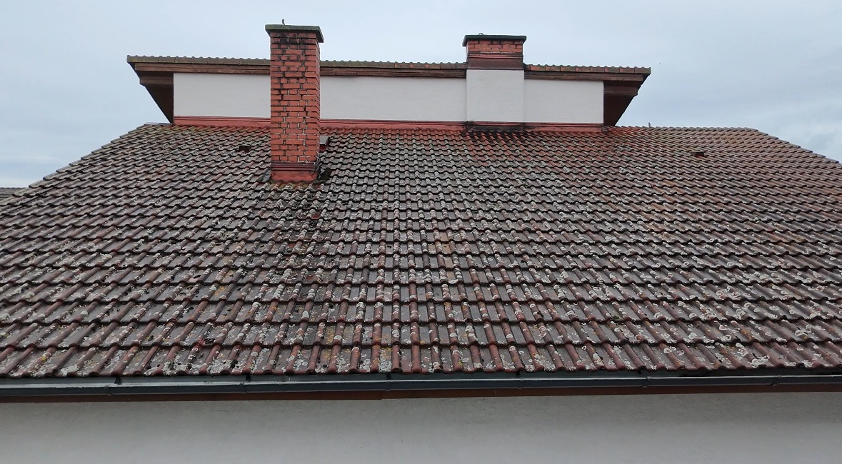 Ciemnobrązowy dach pokryty dachówką ceramiczną, z dwoma kominami. Widoczne ślady mchu i zabrudzeń. Ujęcie z dołu, pochmurne niebo w tle.