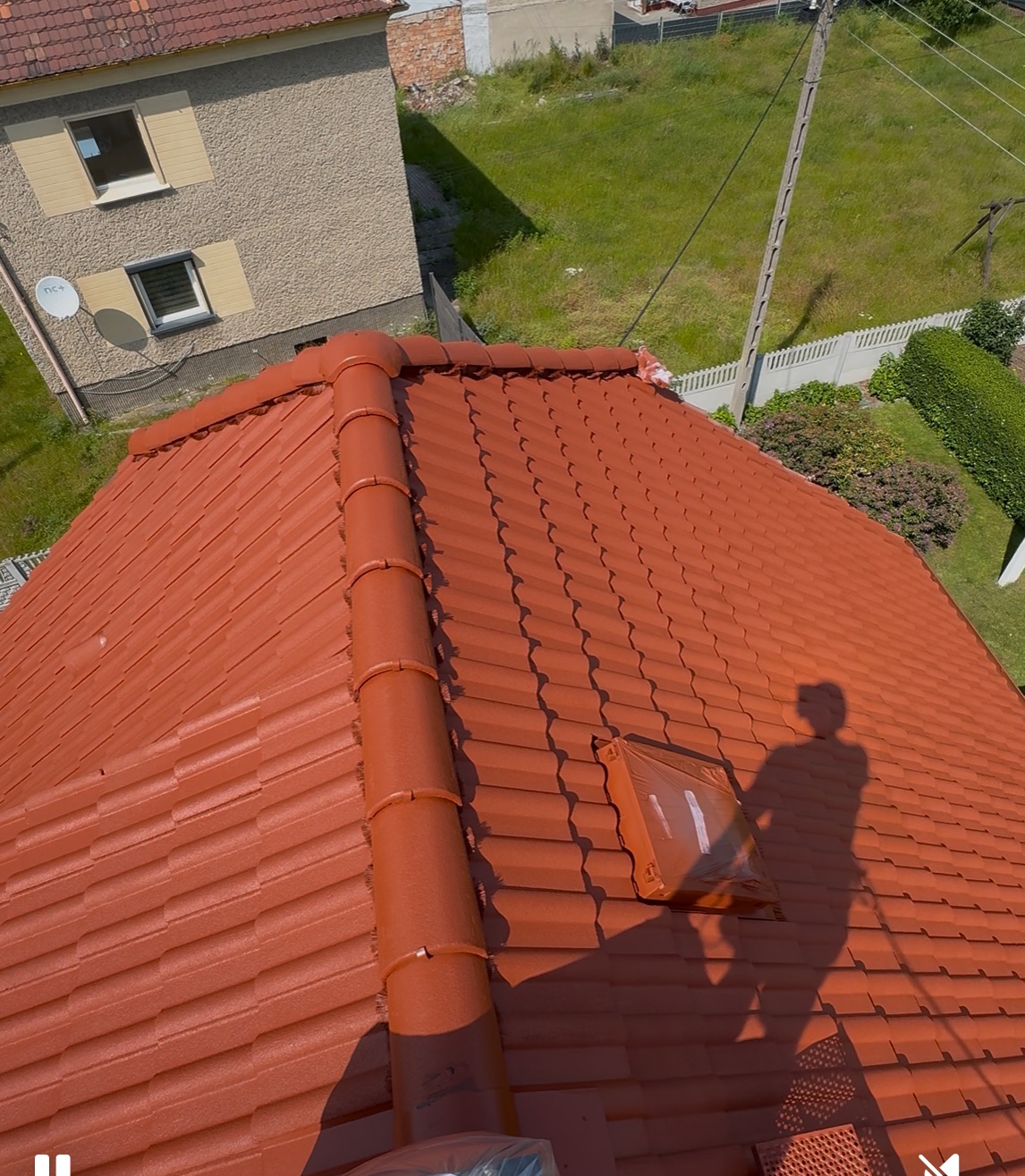 Czerwony dach z dachówki ceramicznej po renowacji, widoczny cień osoby na dachu i okno dachowe. Widok z góry na dom i trawnik w słoneczny dzień.