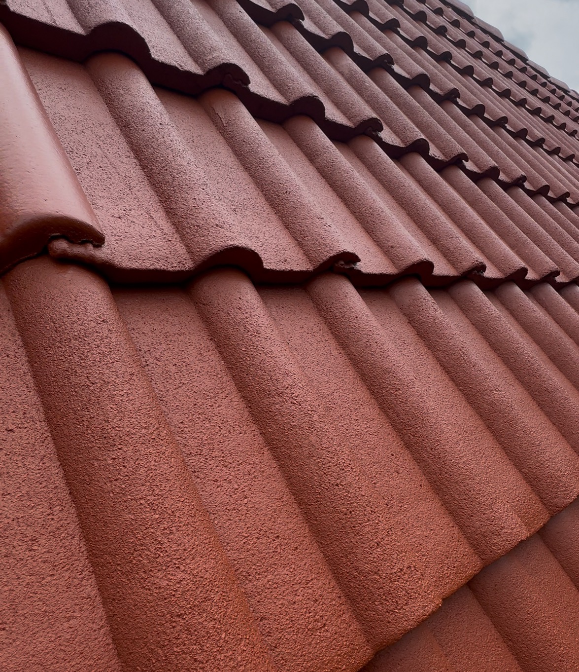 Czerwona, świeżo pomalowana dachówka ceramiczna, widok z dołu pod kątem. Widoczna faktura farby i regularny układ dachówek. Częściowo widoczne niebo w tle.